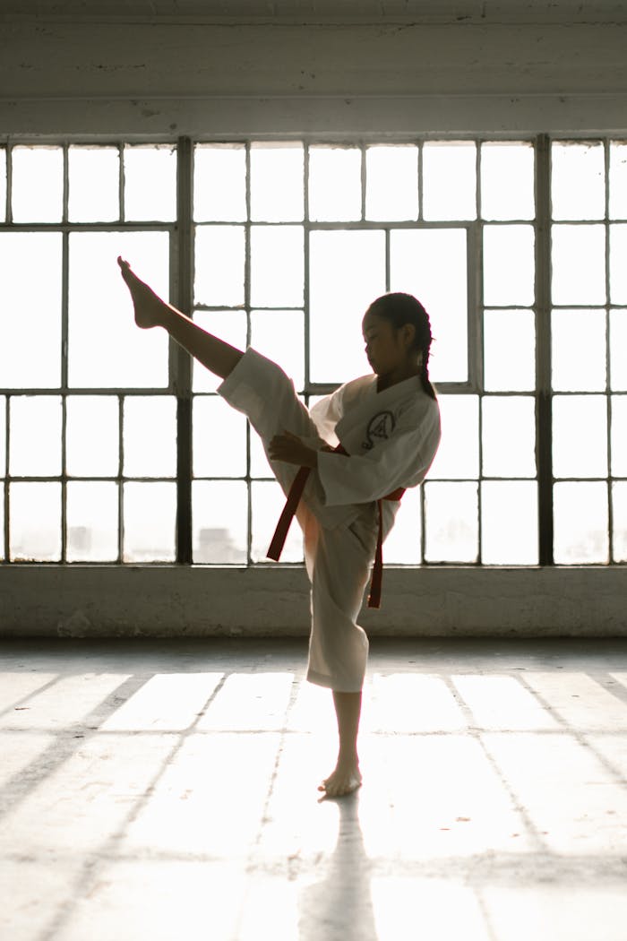 A young girl in a martial arts uniform performing a high kick in a sunlit studio.
